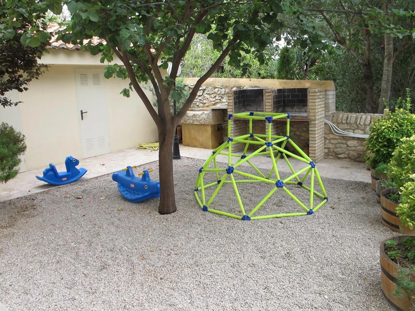 Zona infantil con estructura de juego y balancines bajo un árbol. · Mas dels Estellers