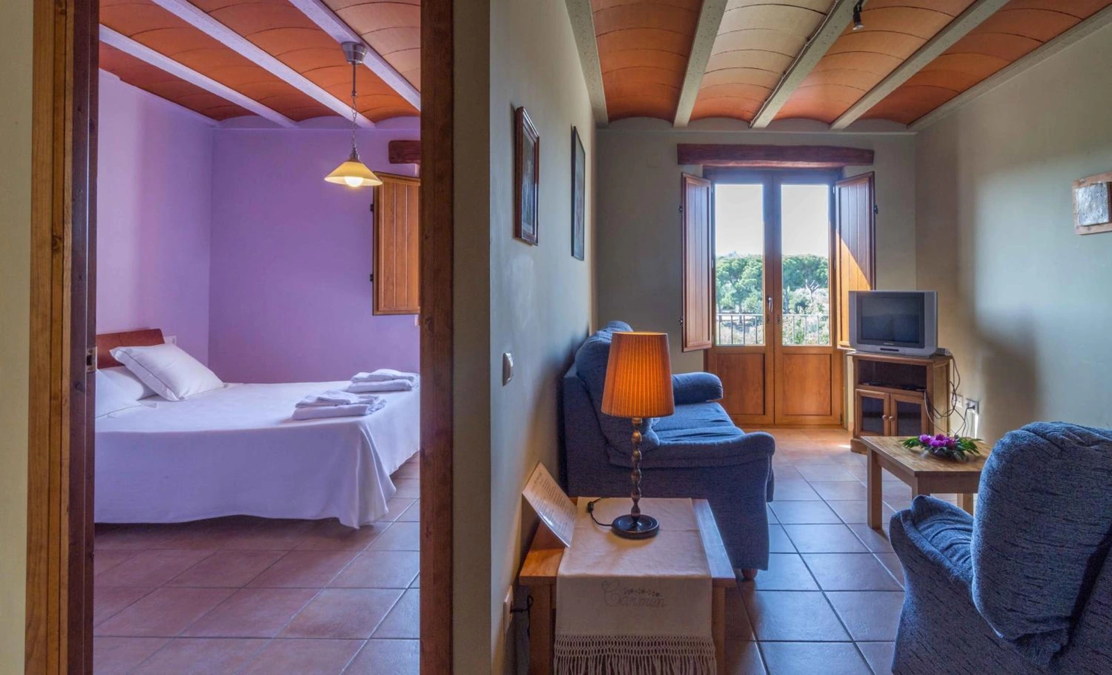 Dormitorio de pared violeta abierto a una zona de estar con sillones azules. · Mas dels Estellers
