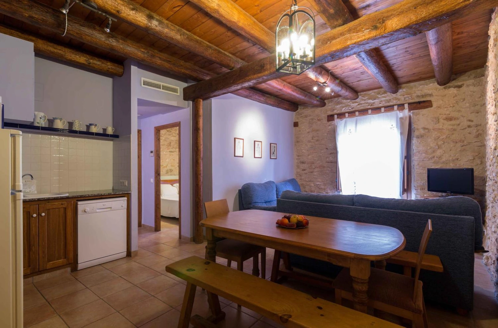 Salón comedor con sofá azul y cocina abierta bajo vigas de madera. · Mas dels Estellers
