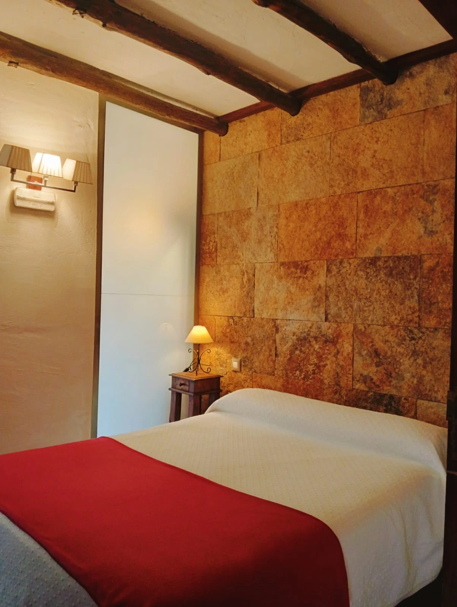 Dormitorio de matrimonio con pared de piedra y colcha roja. · Casa Rural Hidalgo