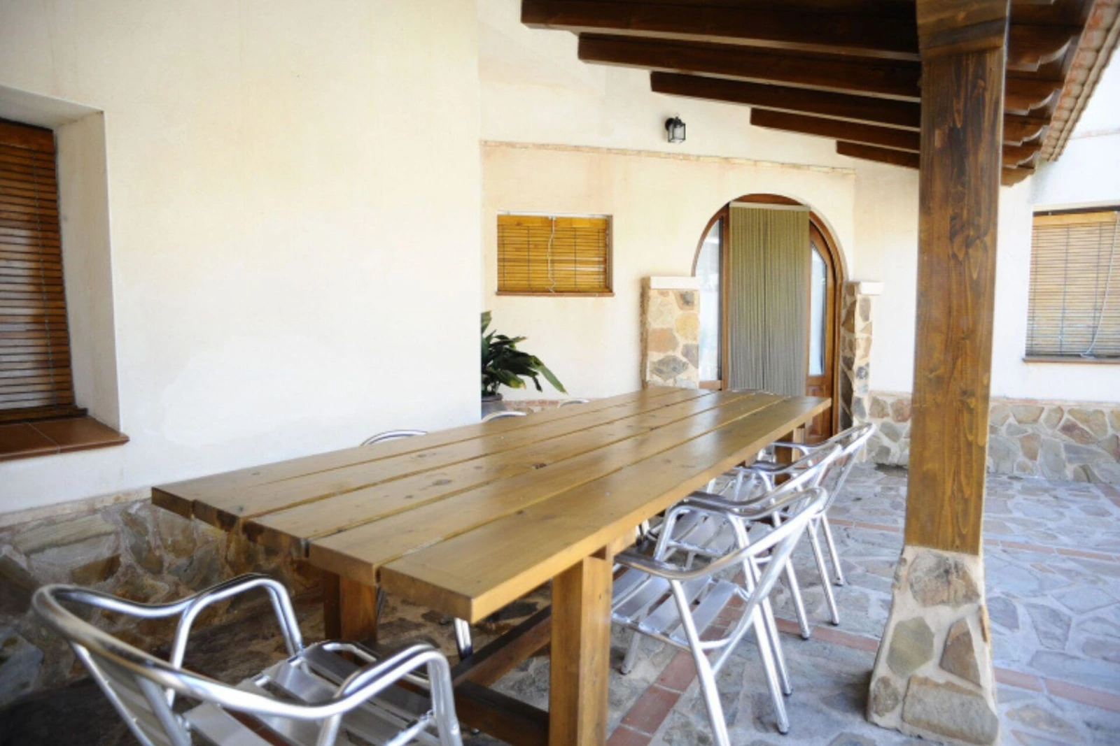 Porche cubierto con una mesa grande y columnas de madera y piedra. · Casa Rural La Plaza