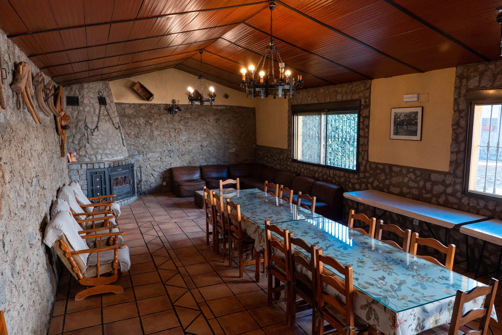 Salón comedor de piedra con mesas largas, chimenea y vigas de madera. · Cortijo Charcollana