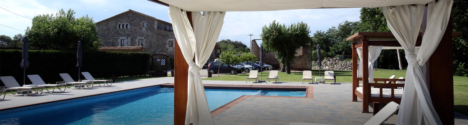 Piscina exterior común de Mas La Casassa, vista entre las cortinas de una pérgola. · Mas La Casassa