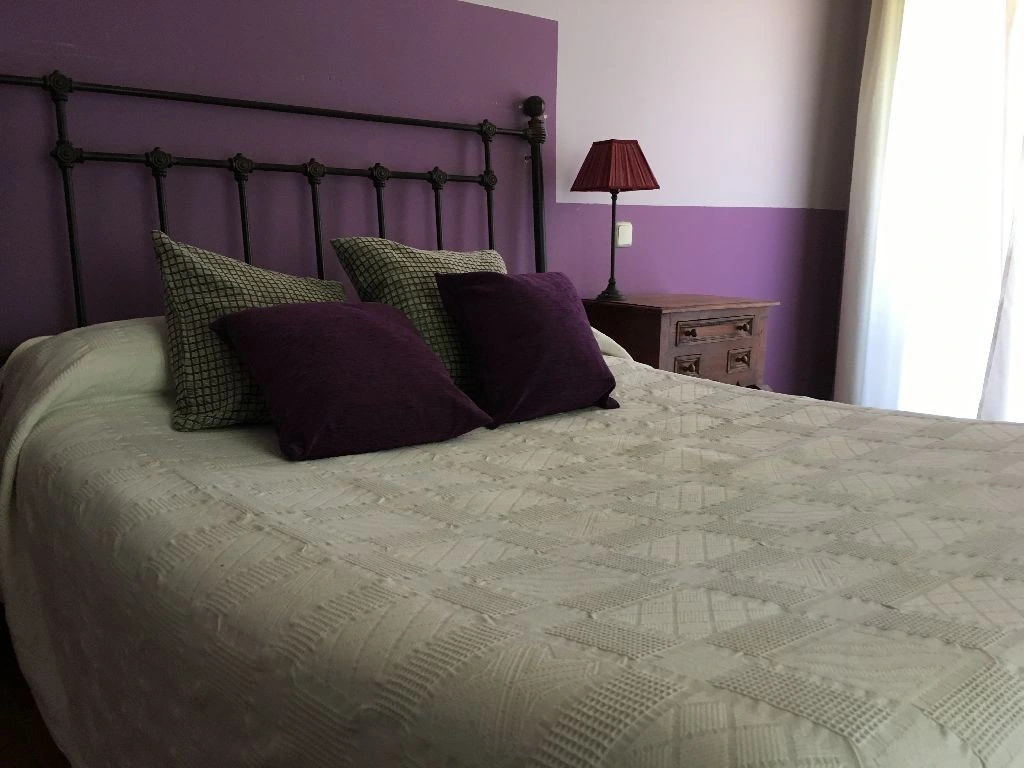 Dormitorio de La Botería con cama de matrimonio, cabecero de hierro y pared violeta · La Botería
